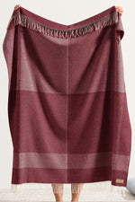 A person holds up a large plaid scarf or shawl in a muted dusty rose or mauve color with fringe trim at the top and bottom. The product is shown against a plain indoor wall, with a small portion of a light cushion visible at the lower right.