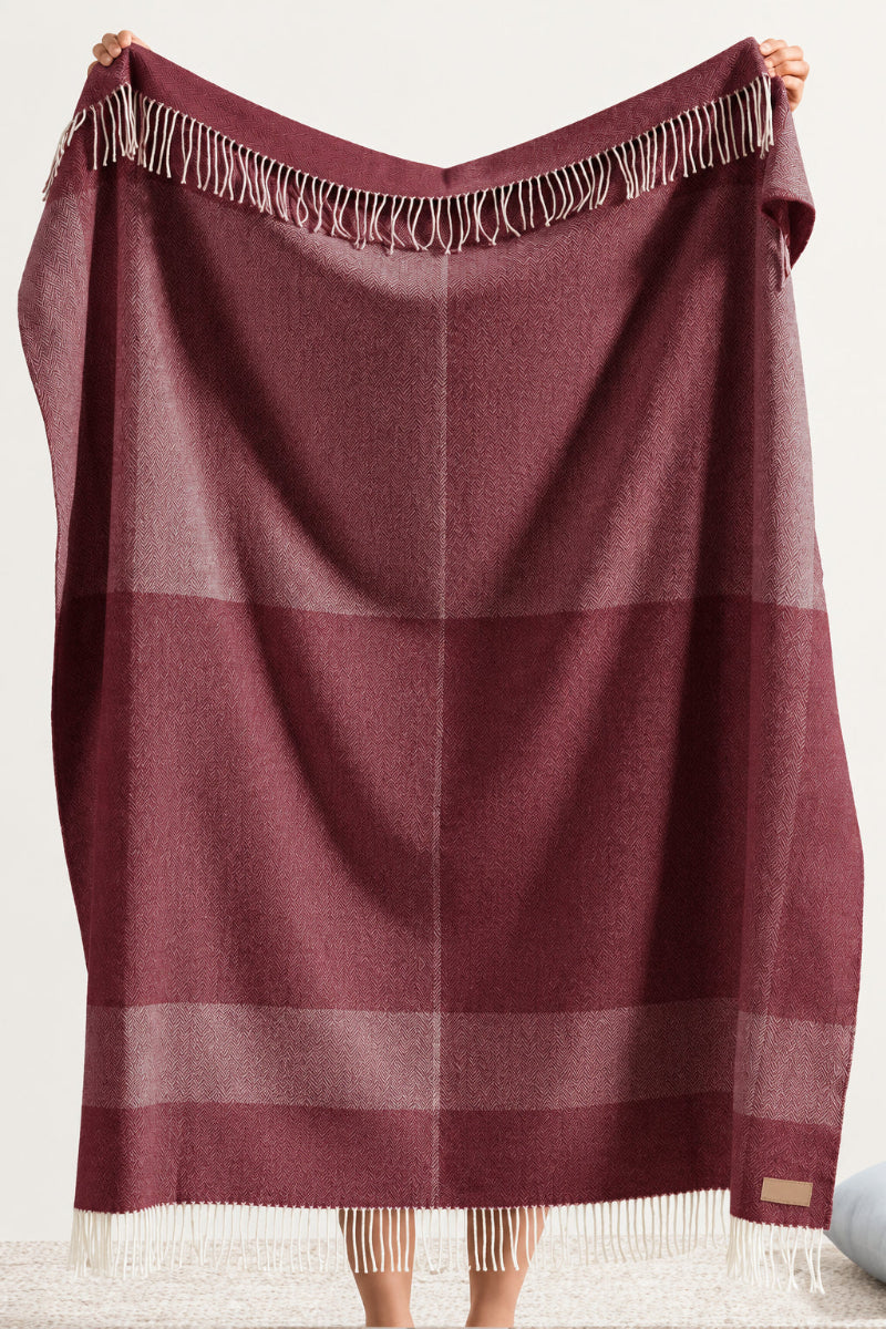 A person holds up a large plaid scarf or shawl in a muted dusty rose or mauve color with fringe trim at the top and bottom. The product is shown against a plain indoor wall, with a small portion of a light cushion visible at the lower right.